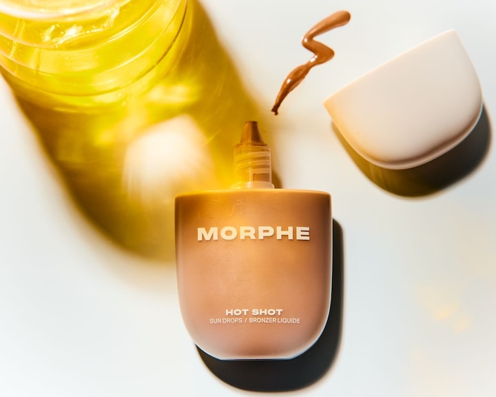 Крупен план на Morphe Hot Shot Sun Drops / течен бронзант в светлокафява бутилка с бял надпис, до него капка от продукта и отворената капачка.