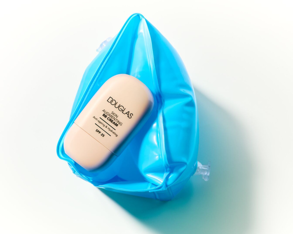 Douglas Skin Augmenting BB крем със SPF 25, в светлопрасковена опаковка, е поставен в яркосин надуваем басейн играчка.